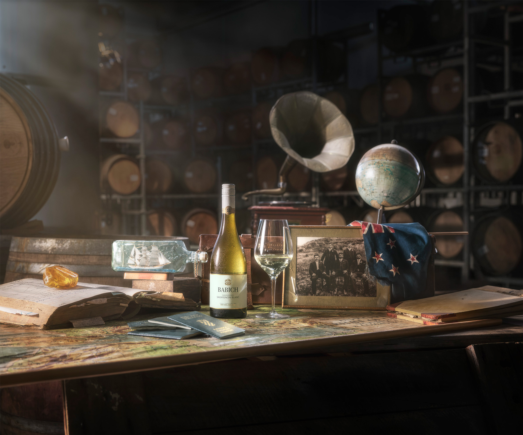Babich Sauvignon Blanc bottle and wine glass displayed on a map-covered table in a cellar, surrounded by vintage objects, a globe, family photo and wine barrels.