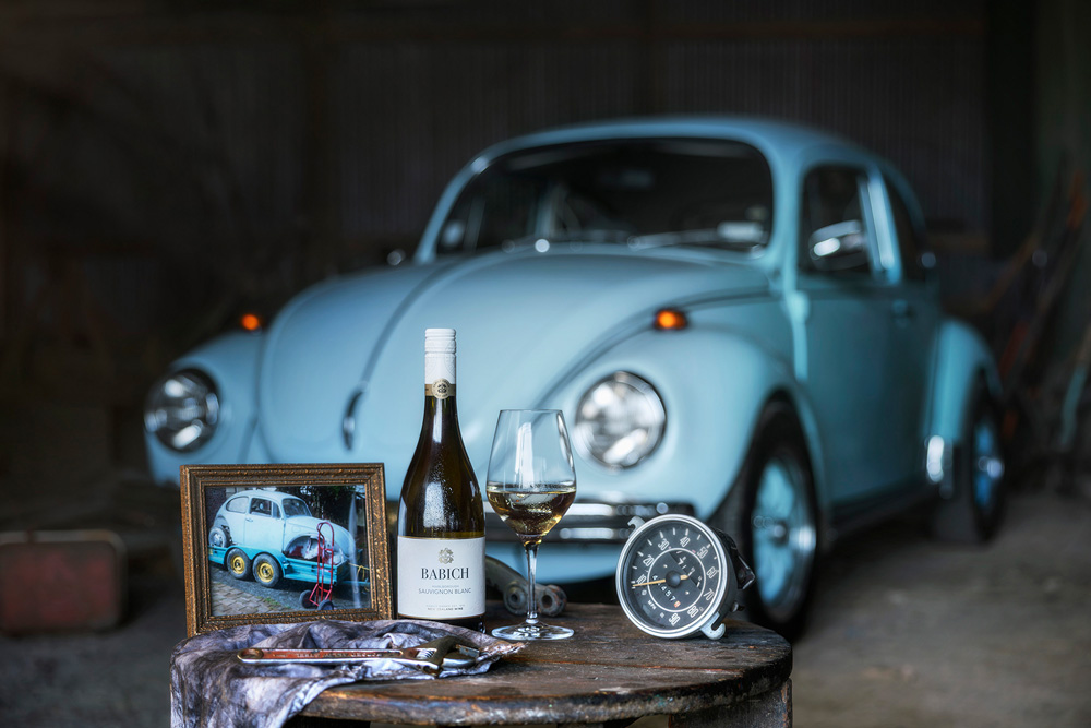 Babich Sauvignon Blanc bottle and wine glass displayed on a small table with vintage car parts and a framed photo, set in front of a blue classic car.