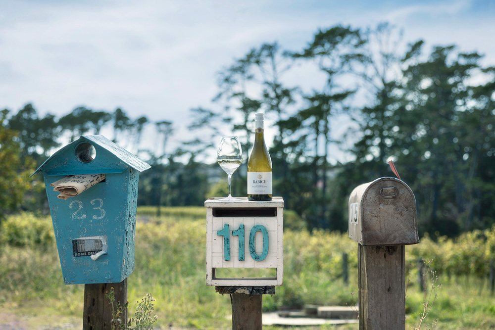 Babich Sauvignon Blanc bottle and wine glass displayed outdoors on a roadside letterbox, with mailboxes and greenery in the background.