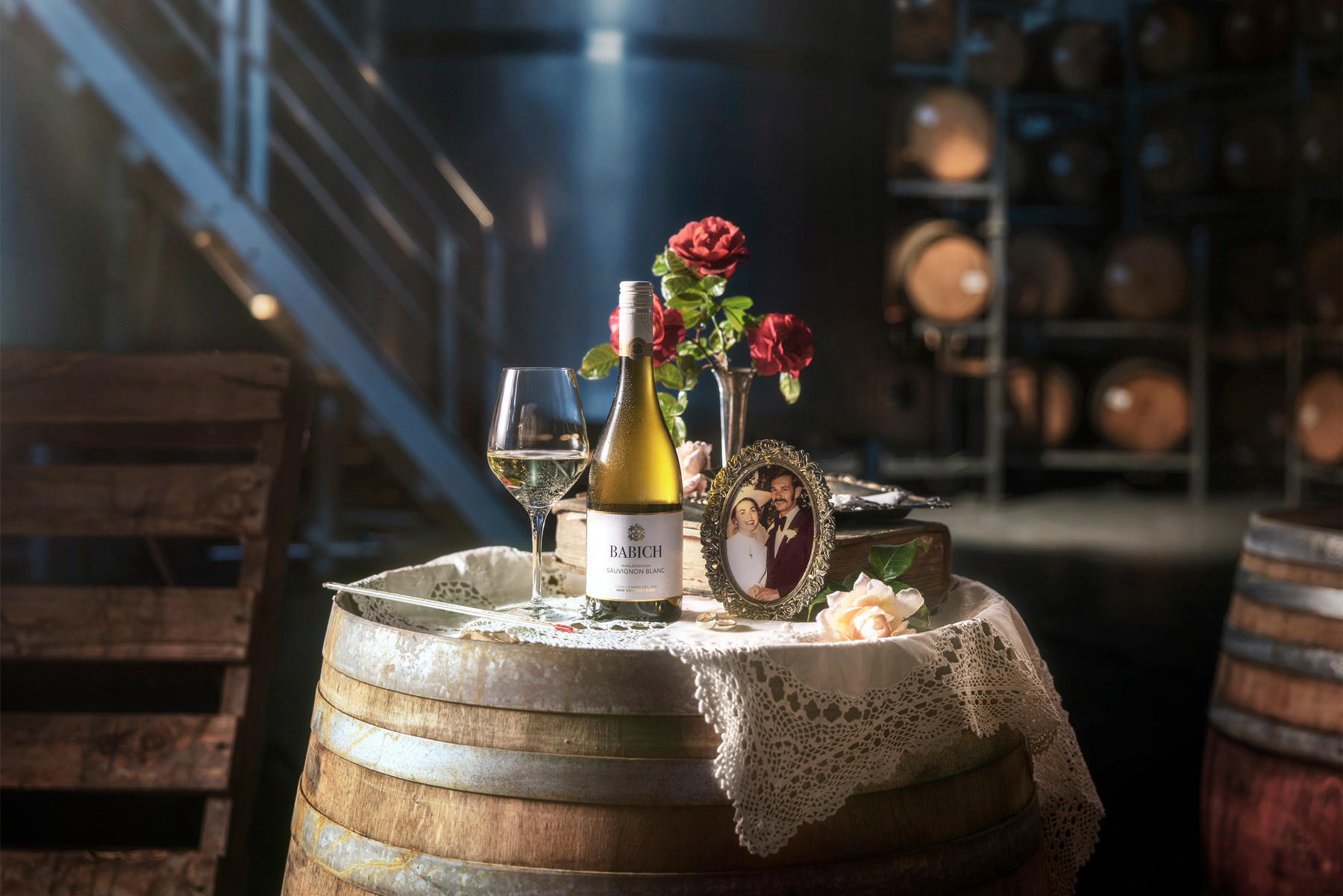 Babich Sauvignon Blanc bottle and wine glass arranged on a lace-covered wine barrel with roses and a framed wedding photo in a dimly lit cellar.