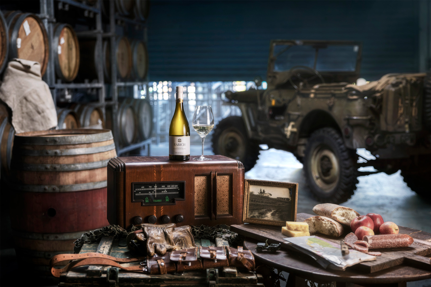 Babich Sauvignon Blanc bottle and wine glass displayed on a vintage radio beside a framed photo, food, maps and military-style memorabilia in a wine cellar.