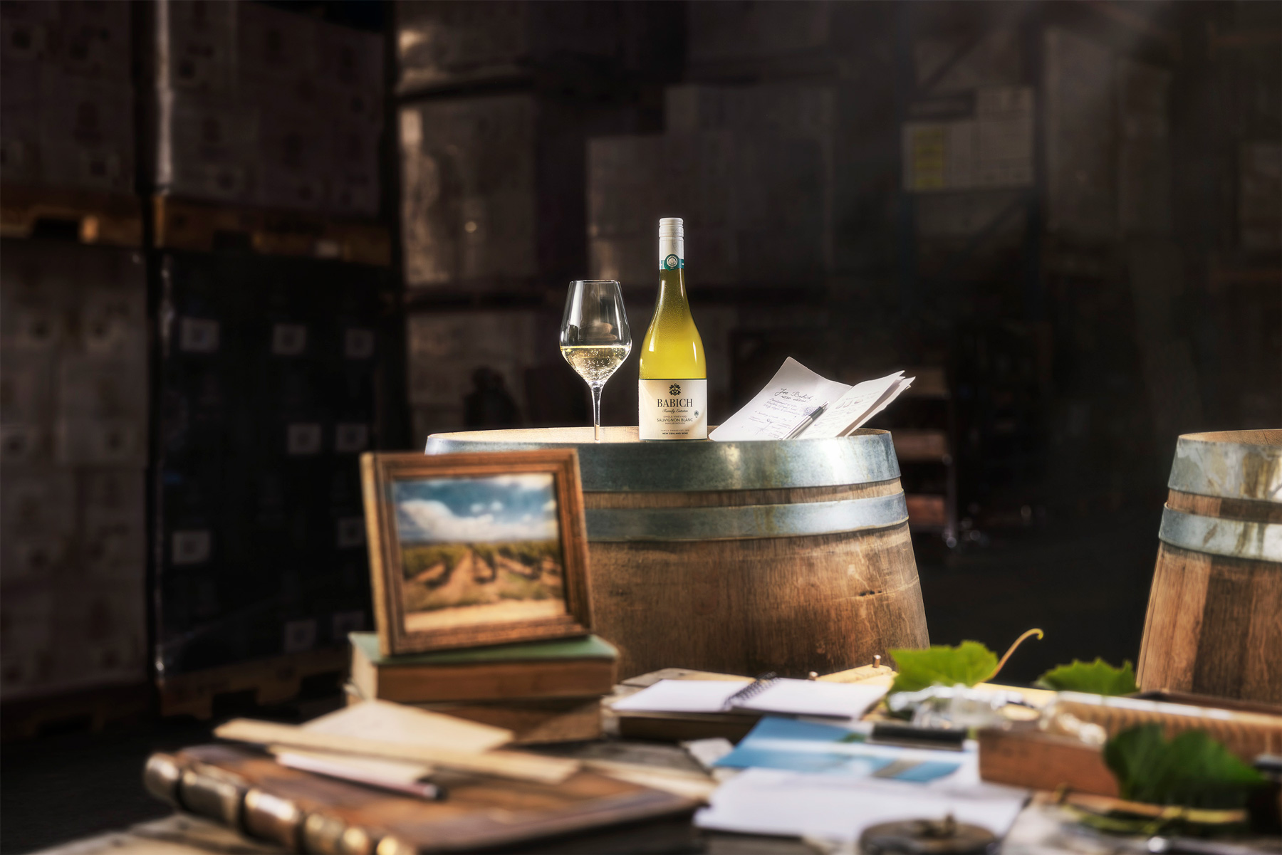 Babich Sauvignon Blanc bottle and wine glass displayed on a wine barrel in a cellar, surrounded by papers, vineyard imagery and grape leaves.
