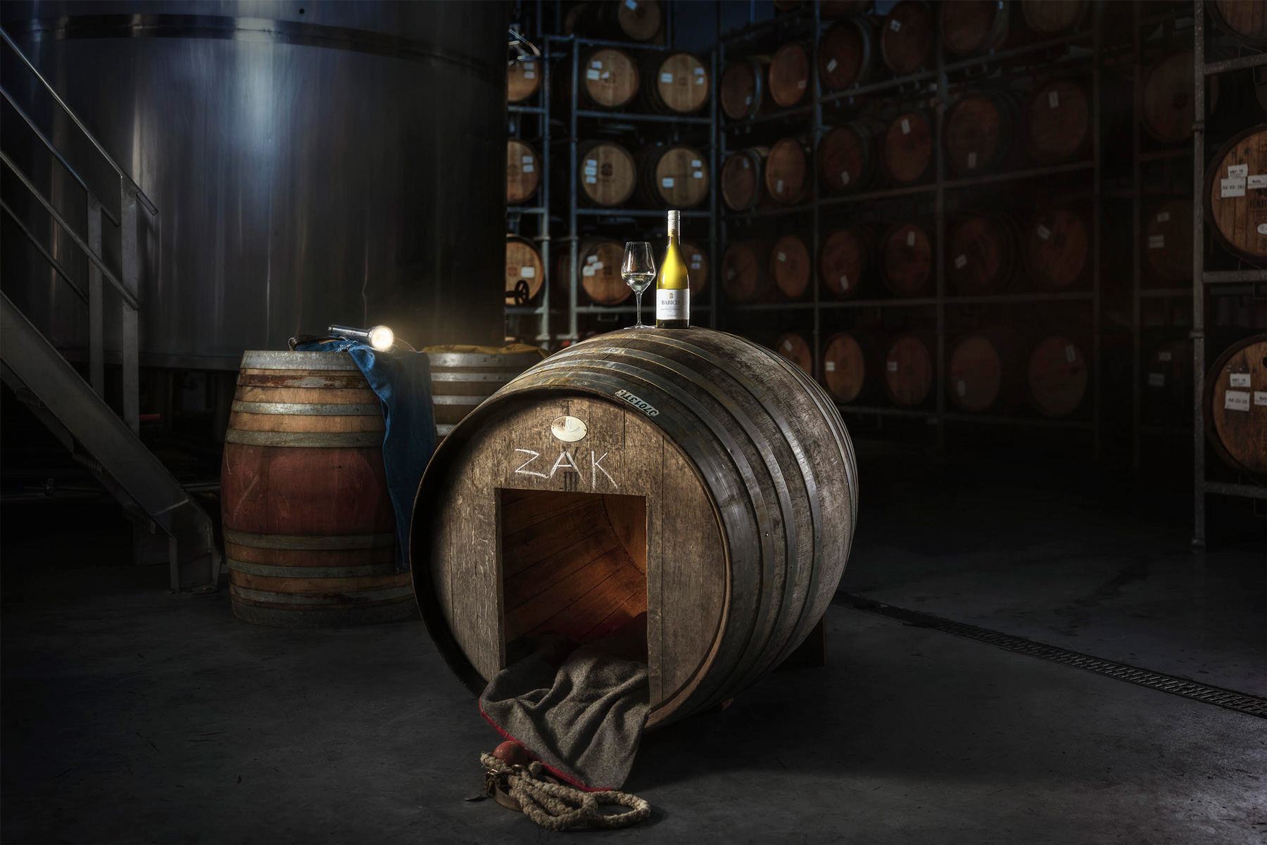 Babich Sauvignon Blanc bottle and wine glass displayed on top of a large wine barrel in a dark cellar, with stacked barrels in the background.