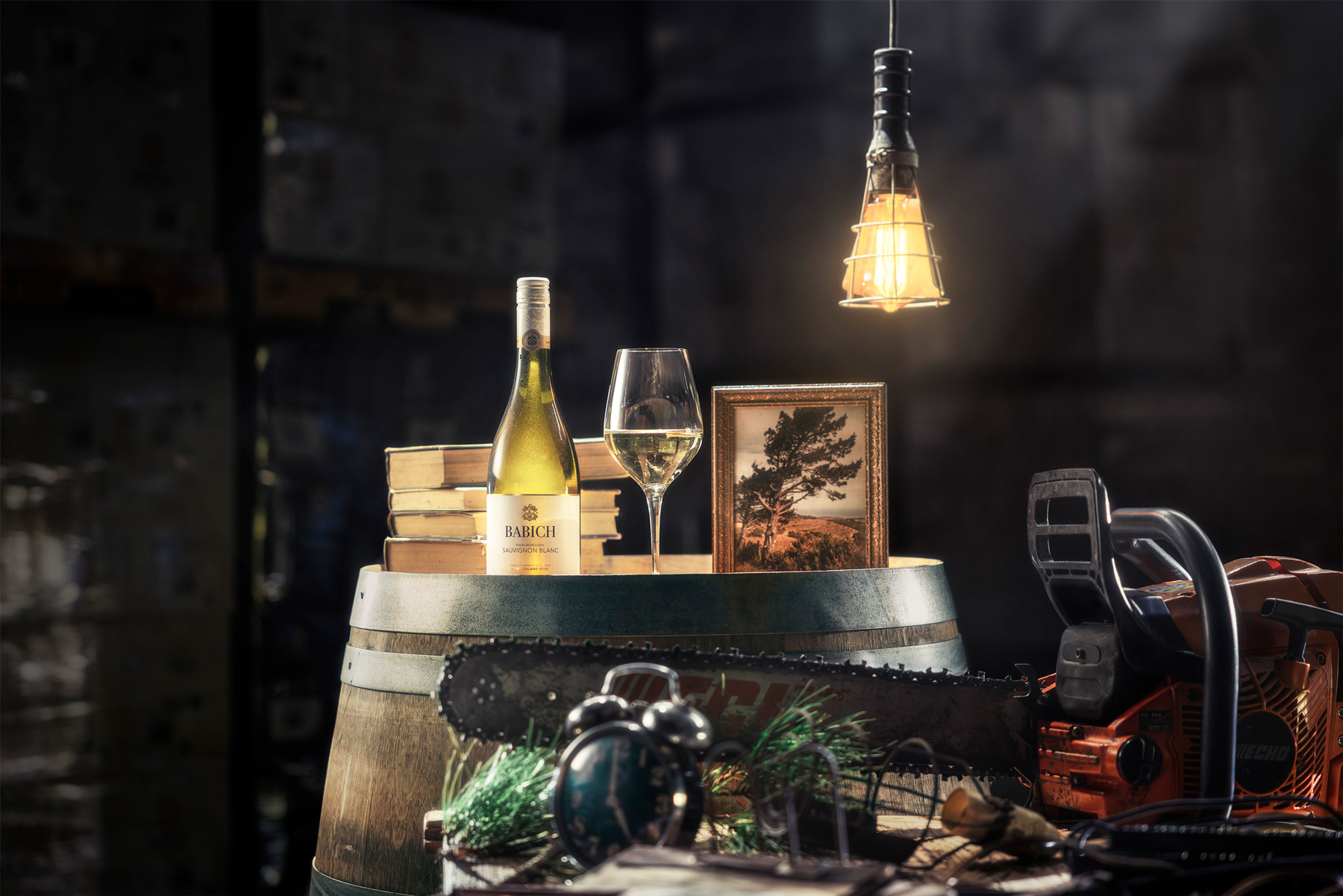 Babich Sauvignon Blanc bottle and wine glass on a barrel with books, a framed photo and work tools, lit by a hanging lamp in a cellar.